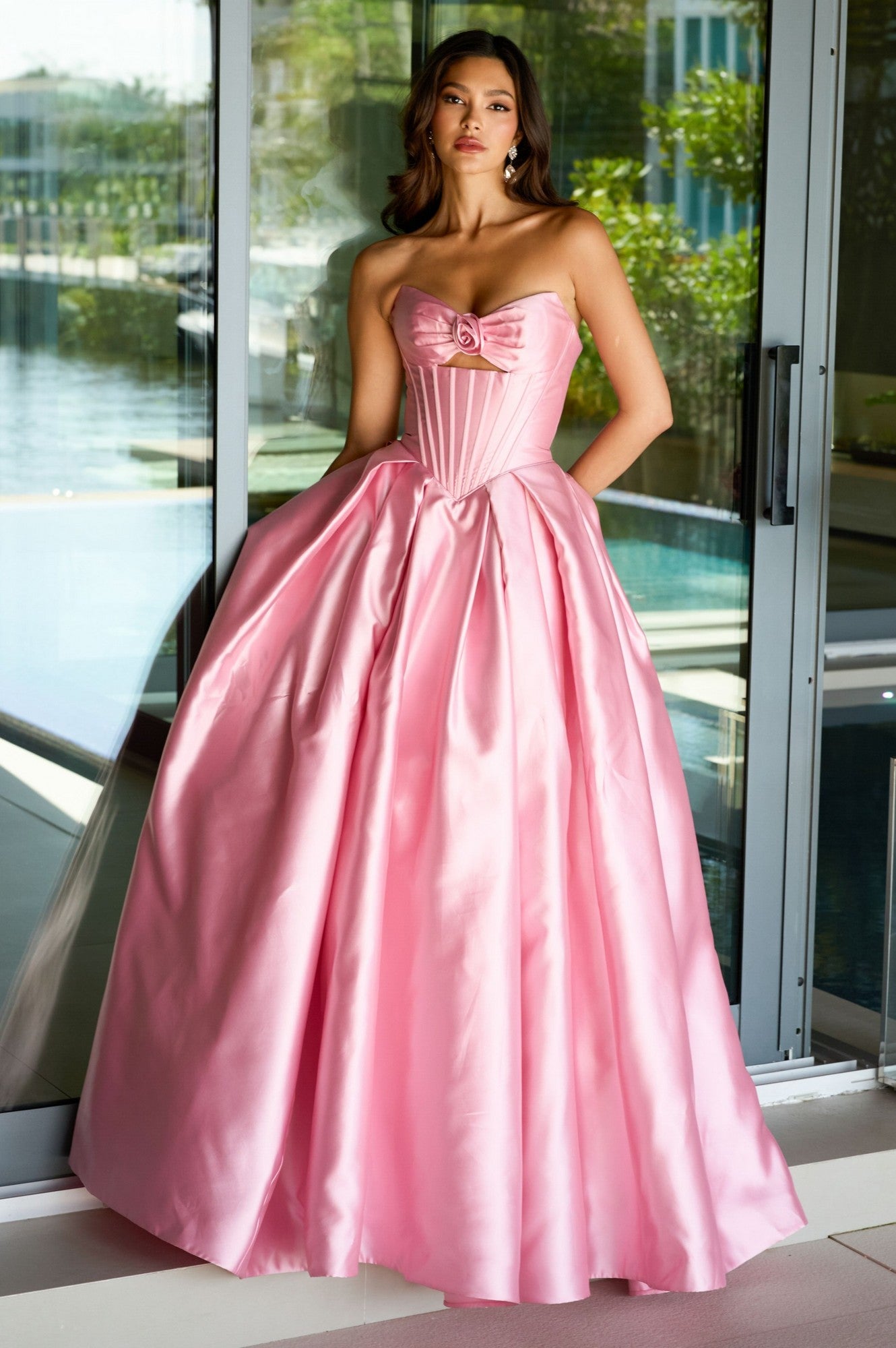 A woman stands indoors in front of glass doors, wearing the Ava Presley 47223 Strapless Satin Prom Ball Gown in blush, featuring a fitted bodice and a full, floor-length skirt.
